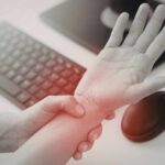 Persona sujetando la muñeca con molestia junto a laptop, señal de dolor por uso repetitivo.