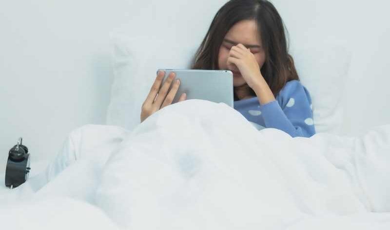 Chica recostada en su cama frotándose los ojos por cansancio provocado por su IPAD
